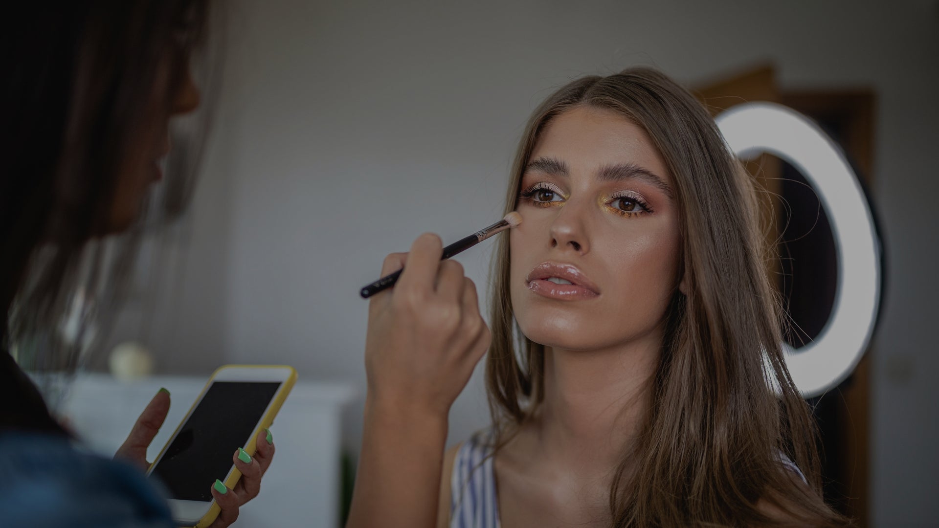 Makeup artist applying makeup to a model using a brush, with a smartphone in hand.