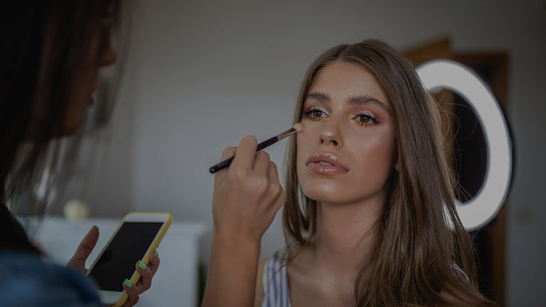Makeup artist applying makeup to a model using a brush, with a smartphone in hand.