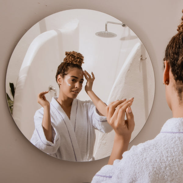 Woman in a white robe applying cream to her face in front of a round mirror.